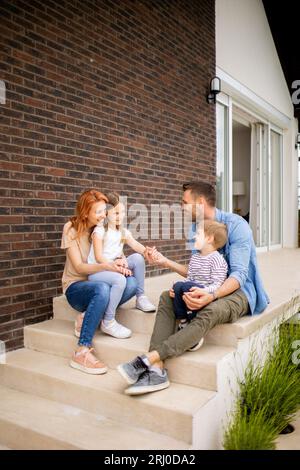 Familie mit einer Mutter, einem Vater, einem Sohn und einer Tochter, die draußen auf den Stufen einer Veranda eines Backsteinhauses sitzen Stockfoto