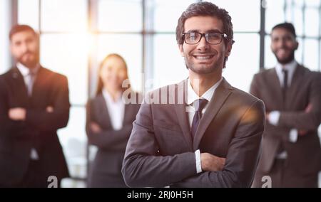 Lächelnder ceo oder professioneller Business Coach, der sich die Kamera im Büro mit einem vielseitigen Team im Hintergrund ansieht, Stockfoto