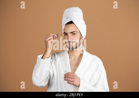 Mann in White Bathrobe mit Gesichtsserum auf Beige-Hintergrund Stockfoto