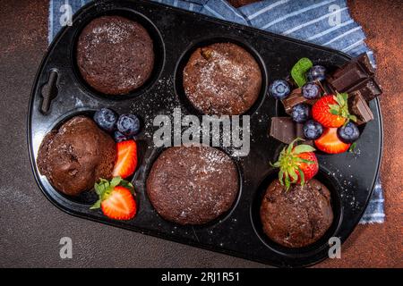 Schokoladen-Muffins mit Beeren. Muffin-Pfanne mit hausgemachten süßen, dunklen Schokoladen-Cupcakes mit Kakaosoße, Zuckerpulver und frischen Erdbeeren, Blaubeeren Stockfoto