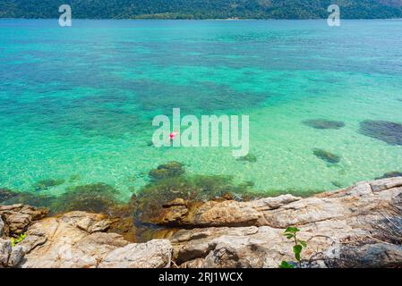 Felsen und türkisfarbenes Wasser mit einem einsamen Schwimmer darin am Sunset Beach in Ko Lipe, Thailand. April Stockfoto