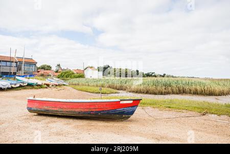 Ein rot-blaues Holzboot, das am Ufer der Brancaster Staithe an der Küste von North Norfolk hinter seinem Seil und Anker abgebildet ist. Stockfoto