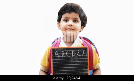 Portrait von glücklichen niedlichen kleinen indischen Jungen in der Schuluniform mit leerem Schiefer vor orangefarbenem Hintergrund, entzückende Grundschule Kind zeigt schwarze Tafel. Stockfoto