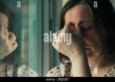 Migräne-Kopfschmerzen, mittelErwachsene kaukasische Frau, die am Wohnzimmerfenster steht und die Stirn drückt, um starke Schmerzen zu stoppen, selektiver Fokus Stockfoto