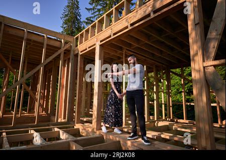 Mann und Frau inspizieren ihre zukünftige Holzrahmenwohnung in der Nähe des Waldes. Ein junges Paar auf der Baustelle am frühen Morgen. Konzept des zeitgenössischen ökologischen Bauens. Stockfoto