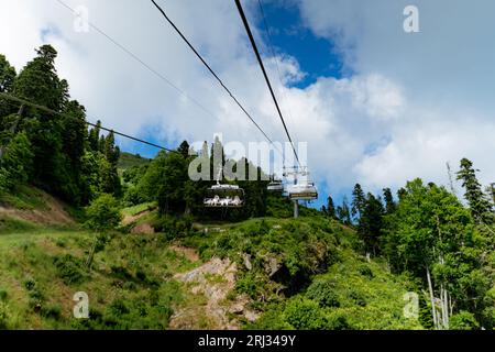 Moderne Seilbahn- oder Seilbahn-Hütten mit Menschen vor dem grünen Tal, blauem Himmel und Berggipfeln. Rosa Khutor, Soschi, Russland Stockfoto