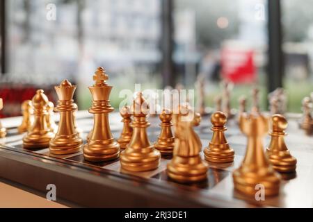 Selektiver Fokus auf die goldenen Stücke des Königs stehen und mit anderen Teilen auf Schachbrett gegen silberne Stücke im unscharfen Innenraum gesetzt. Stockfoto