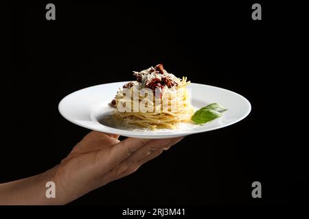 Frau hält Teller mit leckeren Spaghetti mit sonnengetrockneten Tomaten und Parmesankäse auf schwarzem Hintergrund, Nahaufnahme. Exquisite Präsentation von Nudelgerichten Stockfoto