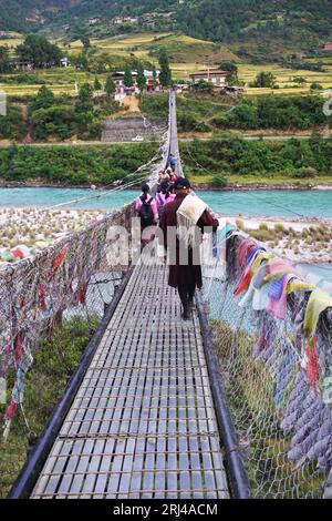 Bhutanese Mann, der traditionelles Gho trägt, geht hinter einer Gruppe von Schulmädchen über die Hängebrücke von Punakha und überquert den Fluss Pho Chhu, Bhutan Stockfoto