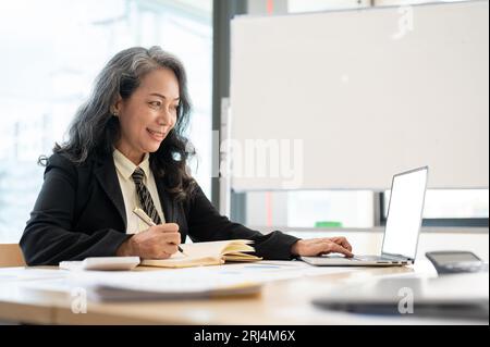 Eine professionelle und elegante Senior-Geschäftsfrau aus Asien oder weibliche Führungskraft, die an ihrem Schreibtisch im Büro auf einem Laptop an ihren geschäftlichen Aufgaben arbeitet. Stockfoto