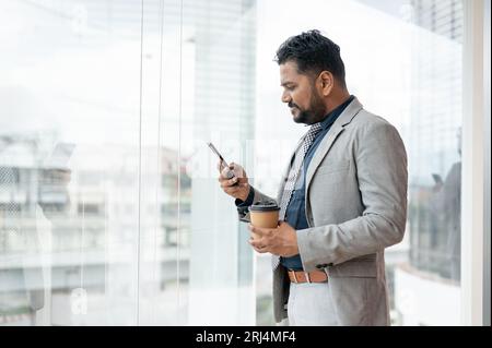 Ein professioneller und erfolgreicher indisch-asiatischer Geschäftsmann oder männlicher CEO in einem formellen Business-Anzug überprüft Nachrichten auf seinem Telefon, während er am W steht Stockfoto