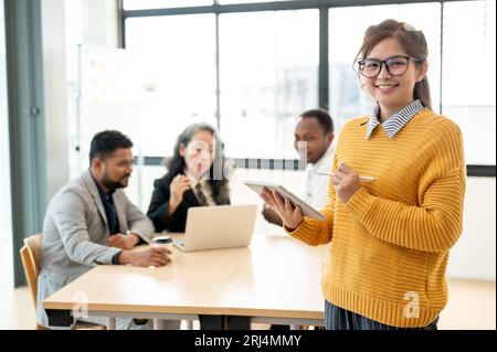 Ein Porträt einer lächelnden und schönen jungen asiatischen Geschäftsfrau in Gläsern, die mit einem Tablet in der Hand in einem Besprechungsraum steht, während ihre Kollegen A Stockfoto
