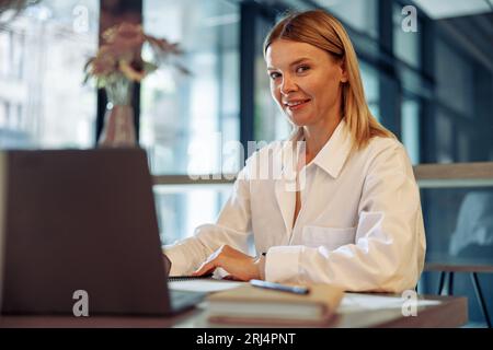 Unternehmerin, die am Laptop arbeitet, während sie in gemütlicher Coworking-Zeit sitzt und die Kamera anschaut Stockfoto