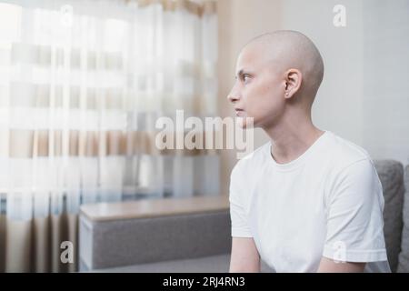 Porträt einer jungen Frau im weißen T-Shirt ohne Haare mit Krebs, schaut traurig zum Fenster Stockfoto