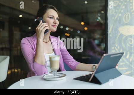Attraktive kaukasische Freiberuflerin, die auf dem Handy spricht, während sie im Café hinter dem Laptop arbeitet Stockfoto