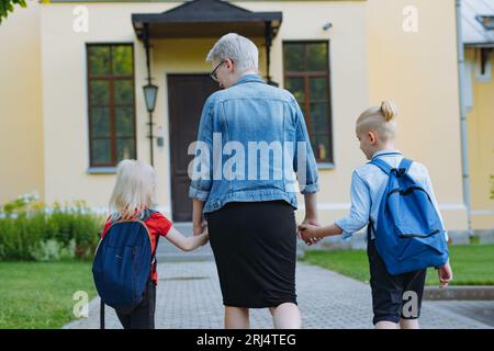 Mutter begleitet Kinder mit Händen zur Schule. Kaukasischer blonder Junge mit im Pferdeschwanz gefesselten Haaren und kleines Mädchen, das mit Mutter zur Schule geht. Bild Stockfoto