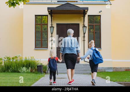 Mutter begleitet Kinder mit Händen zur Schule. Kaukasischer blonder Junge mit im Pferdeschwanz gefesselten Haaren und kleines Mädchen, das mit Mutter zur Schule geht. Bild Stockfoto