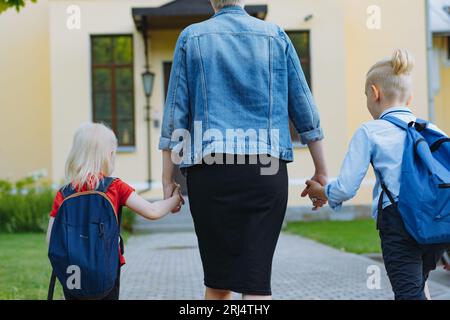 Mutter begleitet Kinder mit Händen zur Schule. Kaukasischer blonder Junge mit im Pferdeschwanz gefesselten Haaren und kleines Mädchen, das mit Mutter zur Schule geht. Bild Stockfoto