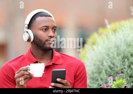 Schwarzer Mann, der Musik über Kopfhörer hört, das Telefon hält und Kaffee in einer Bar trinkt Stockfoto