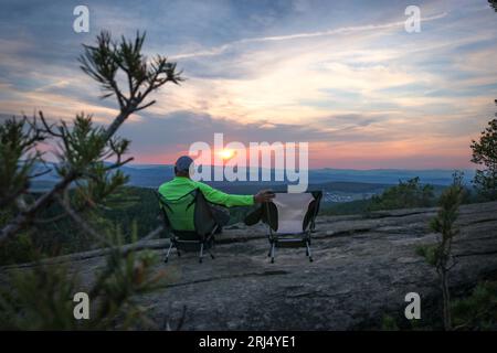 Ein einsamer Mann bewundert den Sonnenuntergang in den Bergen, ein leerer Stuhl steht neben ihm. Das Konzept der Einsamkeit, des Reisens, Zen. Stockfoto