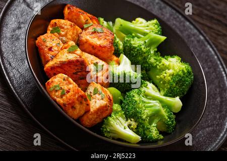 Gegrillte Lachsbrocken mit gedämpften Brokkoliröschen in schwarzer Schüssel auf dunklem Holztisch, Nahaufnahme Stockfoto
