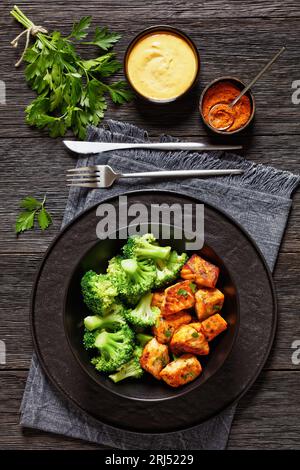 Gebacken im Ofen Lachsbäche mit gedämpften Brokkoliröschen in schwarzer Schüssel auf dunklem Holztisch mit Sriracha Mayo-Sauce, vertikale Ansicht von oben Stockfoto