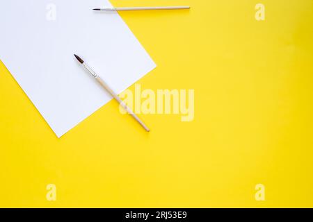 Ein Blatt weißes Papier und Pinsel zum Zeichnen auf gelbem Hintergrund, Draufsicht. Stockfoto