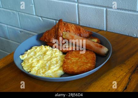 Das Frühstück besteht aus Kartoffelrösti, Croissants, Würstchen und Rührei und wird auf grauen Keramikplatten auf einem Holztisch serviert. Nahrung Photogra Stockfoto