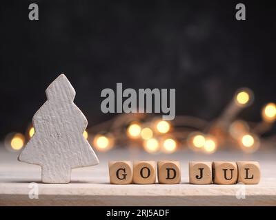 Kleine Holzblöcke mit God Jul und weißem hölzernem Weihnachtsbaum, verschwommene Lichter im Hintergrund Stockfoto