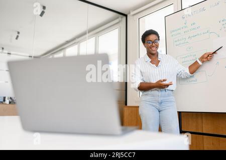 Selbstbewusste junge afrikanerin führt Webinar, Business Training online durch. Der Online-Coach steht in der Nähe des Flipcharts vor dem Laptop und erklärt dem Online-Publikum etwas Stockfoto