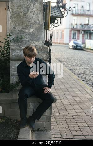 Blonder Mann in einem schwarzen Hemd sitzt mit einem Telefon in den Händen Stockfoto