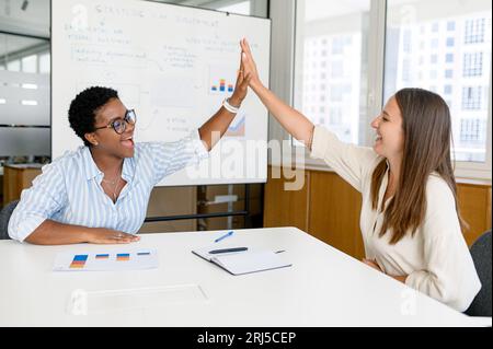 Zwei unterschiedliche weibliche Mitarbeiter in eleganter Freizeitkleidung, die sich im modernen Tagungsraum in Innenräumen hohe fünf leisten, um die erfolgreiche Zusammenarbeit zu feiern, unterstützen sich Kollegen gegenseitig Stockfoto