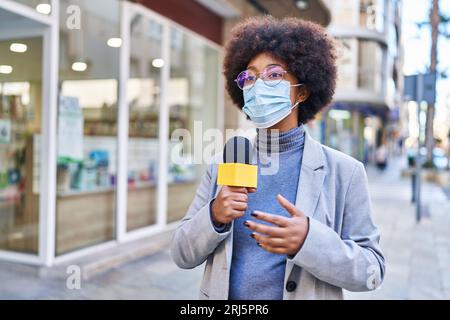 afroamerikanische Reporterin trägt eine medizinische Maske und benutzt ein Mikrofon, das auf der Straße arbeitet Stockfoto