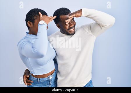 Junge afroamerikanische Paare, die über blauem Hintergrund stehen, sind sehr glücklich und lächelnd, wenn sie weit weg mit der Hand über dem Kopf schauen. Konzept wird gesucht. Stockfoto