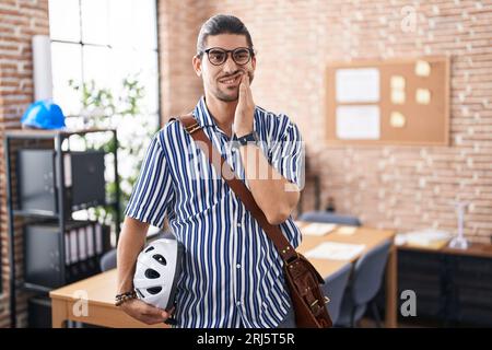 Hispanischer Mann mit langem Haar, der im Büro arbeitet und den Fahrradhelm hält, der den Mund mit der Hand berührt, mit schmerzhaftem Ausdruck wegen Zahnschmerzen oder Zahnschmerzen Stockfoto