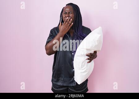Junge afrikanerin, die Pijama trägt und Kissen umarmt, langweilig gähnt, müde, den Mund mit der Hand bedeckt. Ruhelosigkeit und Schläfrigkeit. Stockfoto