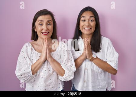 Hispanische Mutter und Tochter beten mit Händen zusammen und bitten um Vergebung, und lächeln selbstbewusst. Stockfoto