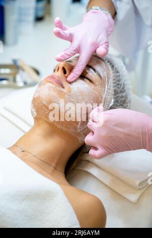 Vor der PRP-ttherapie trägt der Kosmetologe Film auf die Creme mit Anästhesie auf das Gesicht der Frau auf. Stockfoto