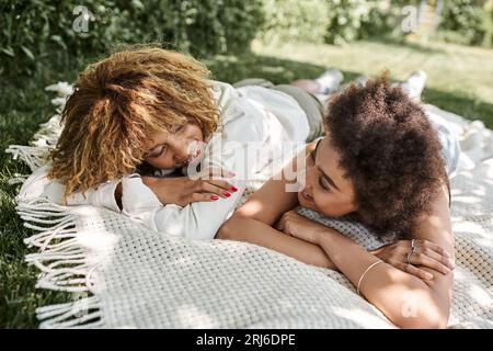 Junge afroamerikanische Freundinnen, die sich auf einer Decke im Park entspannen und sich gegenseitig anlächeln Stockfoto