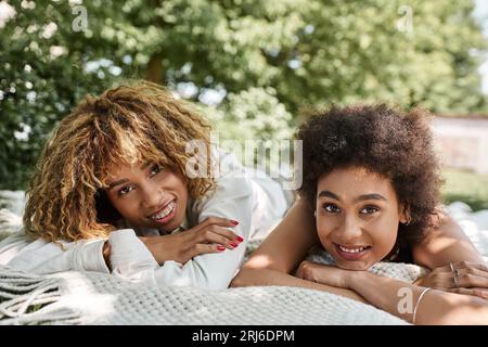 Junge afroamerikanische Freundinnen, die im Park auf einer Decke liegen und vor der Kamera lächeln, Sommerferien Stockfoto