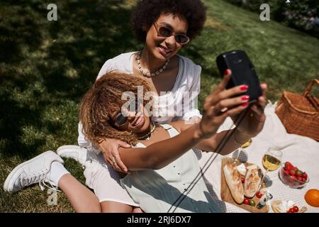 Fröhliche afroamerikanische Frau in Sonnenbrille, die Selfie mit ihrer Freundin im Sommerpark macht Stockfoto