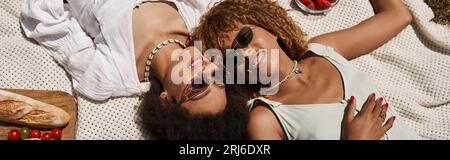 Fröhliche afroamerikanische Freunde, die in der Nähe von Brot, frischem Gemüse, Sommerpicknick, Blick von oben, Banner liegen Stockfoto