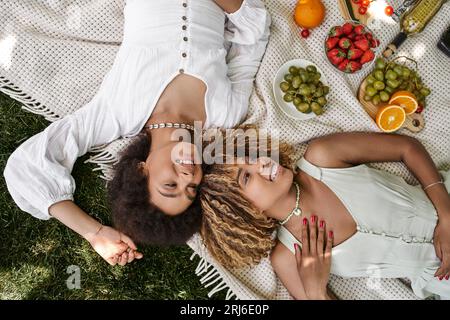 Junge afroamerikanische Freundinnen, die auf einer Decke liegen, Obst und Gemüse, Sommerpicknick, Blick von oben Stockfoto