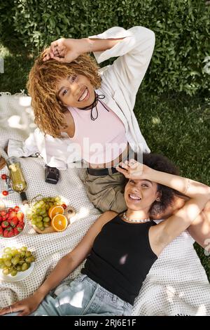 Top-Blick auf sorgenfreie afroamerikanische Freundinnen in der Nähe von Wein und frischem Obst beim Sommer-Picknick Stockfoto