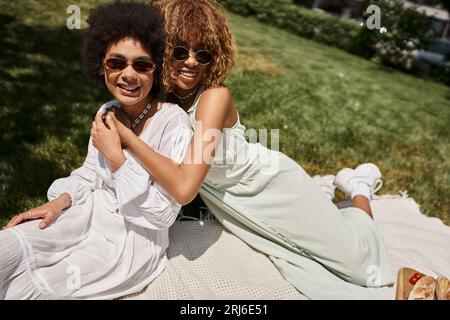 Sorglose afroamerikanische Freundinnen in Sonnenbrillen, die sich umarmen und die Kamera betrachten, Sommerpicknick Stockfoto