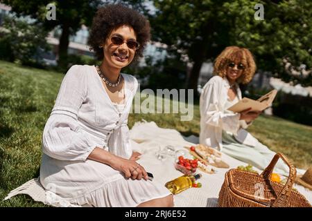 afroamerikanische Frau mit Smartphone, die in der Nähe ihrer Freundin auf die Kamera schaut, Sommerpicknick Stockfoto