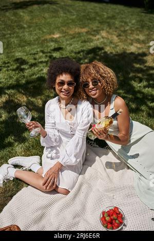 Fröhliche afroamerikanische Freundinnen, weiße Kleider, Weinflasche, Gläser, Sommer-Picknick Stockfoto