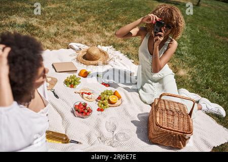 Junge afroamerikanische Frau fotografiert Freundin auf Vintage-Kamera, Picknick im Sommerpark Stockfoto