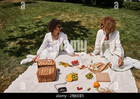 Positive afroamerikanische Freundinnen in der Nähe von Obst, Gemüse auf Decke, Sommerpicknick Stockfoto