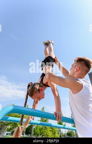 Mädchen, das in parallelen Stangen Liegestütze ausübt. Der Personal Trainer hilft einem Mädchen, einen Handstand auf den unebenen Stangen zu halten. Stockfoto